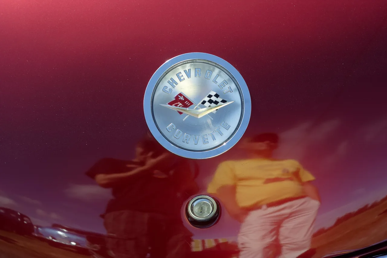 Chrome Chevrolet Corvette nose emblem on a maroon C1, with two people reflected in the paint